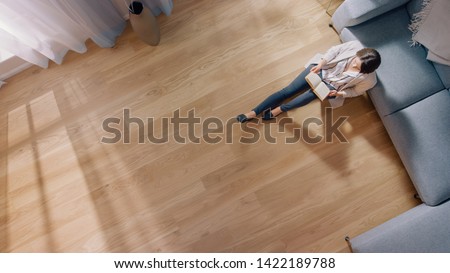 Young Woman is Sitting on a Floor and Reading a Book. Cozy Living Room with Modern Interior, Grey Sofa and Wooden Flooring. Top View Camera Shot.