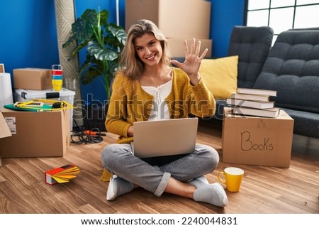 Young woman sitting on the floor at new home using laptop showing and pointing up with fingers number five while smiling confident and happy. 
