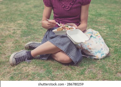 A Young Woman Is Sitting On The Grass In A Park And Is Eating Falafel