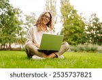 Young Woman sitting on green grass ground, working on laptop in city park.  Mobile Office. Concept for education, business, blog or freelance.