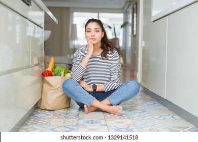 Young woman sitting on the kitchen floor with a paper bag full of fresh groceries thinking looking tired and bored with depression problems with crossed arms. - Powered by Shutterstock