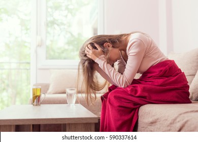 Young woman sitting on a living room couch, covered with a blanket, holding her head, having a splitting headache and suffering enormous pain