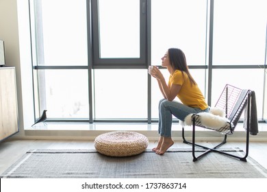 Young Woman Sitting On Modern Chair In Front Of Window Relaxing In Her Living Room And Drinking Coffee Or Tea