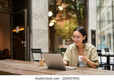 Young woman sitting on online meeting in outdoor cafe, talking to laptop camera, explaining something, drinking coffee. - Powered by Shutterstock