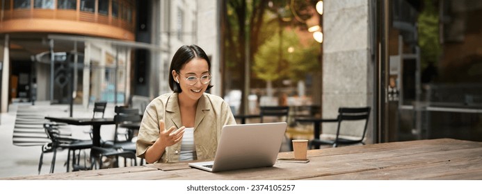 Young woman sitting on online meeting in outdoor cafe, talking to laptop camera, explaining something, drinking coffee. - Powered by Shutterstock