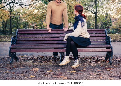 A Young Woman Is Sitting On A Park Bench And Is Talking To A Man Who Is Standing Behind Her