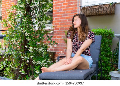 Young Woman Sitting On Patio Lounge Chair In Outdoor Spring Garden In Backyard Of Home With Green Plants And White Flowers