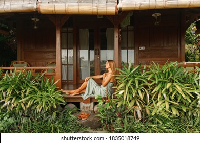 Young Woman In Sitting On A Porch Or Veranda Of Wooden Summer Cottage Or Bungalow. Tropical Summer Vacation.