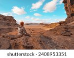 Young woman sitting on rocky ground in desert landscape, small vehicle far distance. View from behind. Wadi Rum, Jordan