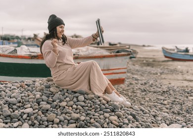 Young woman is sitting on a rocky beach, taking a selfie with her tablet in front of fishing boats - Powered by Shutterstock