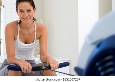 Young Woman Sitting On A Rowing Machine In Her Home And Smiling At The Camera While Holding The Bar In Her Hand