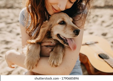 Young woman sitting on the sand on a beach, hugging her little dog, cocker spaniel breed puppy.