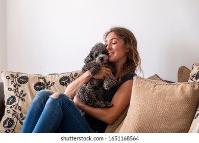 Young Woman Sitting On Sofa Cuddling Cockapoo Puppy