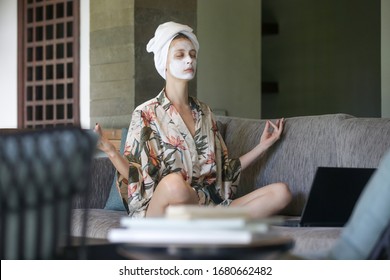 Young woman sitting on sofa with face beauty mask in front of laptop and doing yoga or meditation. Cozy time at home. Self time and home spa concept. Coronavirus outbreak and self-quarantining concept