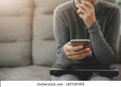 Young Woman Sitting On Sofa, Holding And Looking The Phone Worried Or Thoughtful.Thinking About Solution For The Problem.Work At Home Advice To Stop Coronavirus COVID-19 Spreading.