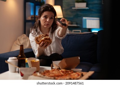 Young Woman Sitting On Sofa Eating Double Cheeseburger Holding Remote Turning Up Volume On Television Watching Evening News. Person Changing Tv Channels While Eating Takeaway Hamburger.