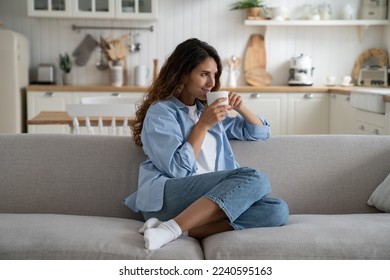 Young woman sitting on sofa at home with cup tea and looking aside with smile on face, starting day with coffee. Calm contented female holding mug enjoying warm beverage, resting relaxing on weekend - Powered by Shutterstock