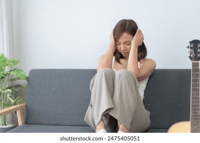 A young woman sitting on a sofa covering her ears, displaying stress or anxiety, with a guitar and plant in the background. - Powered by Shutterstock