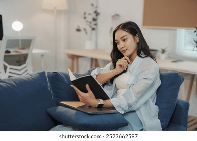 Young woman is sitting on a sofa in her living room, reading a book and enjoying a relaxing evening at home - Powered by Shutterstock