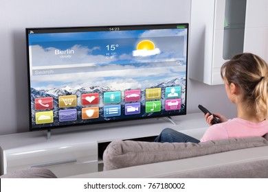 Young Woman Sitting On Sofa Using Remote Control In Front Of Television