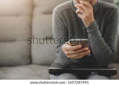 Young woman sitting on sofa, holding and looking the phone worried or thoughtful.Thinking about solution for the problem.Work at home advice to stop coronavirus COVID-19 spreading.
