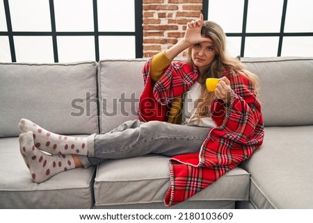 Young woman sitting on the sofa drinking a coffee at home making fun of people with fingers on forehead doing loser gesture mocking and insulting. 