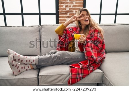 Young woman sitting on the sofa drinking a coffee at home doing peace symbol with fingers over face, smiling cheerful showing victory 
