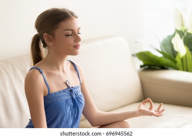 Young woman sitting on soft sofa in lotus position. Calm lady meditating on couch with crossed legs, practicing yoga at home in the morning, concentrating on positive thoughts, struggling with stress