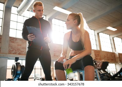 Young Woman Sitting On A Stool And Discussing Exercise Plan With Personal Trainer. Trainer Making An Fitness Plan For Young Female Client At Gym.