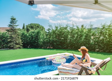 Young Woman Sitting On A Sun Lounger In A Bikini, By The Pool Looking At Her Smartphone. Girl Enjoying Her Summer Holiday. Concept Leisure And Travel. Sunlight, Outside A Garden.