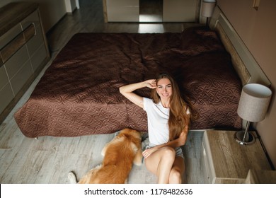 Young Woman Sitting On Te Floor In Her Bedroom And Smiling, Dog Hiding Under Bed