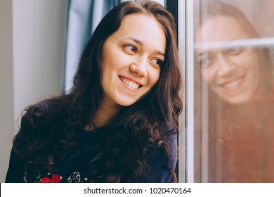 Young Woman Sitting On Window Sill Looking Out The Window And Smiling. Reflection Of The Face In The Window.