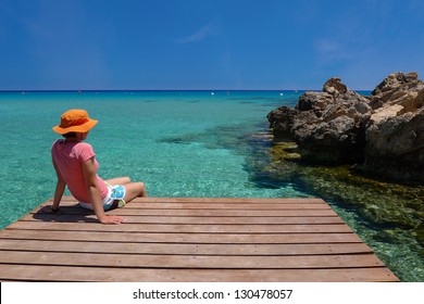 Young Woman Sitting On Wooden Jetty And Looking At Beautiful Crystal Clear Sea Water , Konnos Bay Beach, Protaras, Cyprus