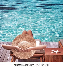 Young Woman Sitting On Wooden Pier On The Sea Wearing Hat And Taking Sun Bath, Enjoying Perfect Summer Day, Traveling And Luxury Vacation Concept