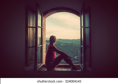 Young woman sitting in an open old window looking on the landscape of Tuscany, Italy. Conceptual romantic, dreaming, hope, travel.