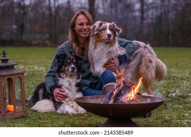 Young woman is sitting outside in the woods with her two Australian Shepherd dogs. Snow on the grass, twilight by the campfire. Cuddles and companion animals - Powered by Shutterstock