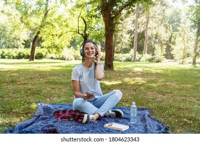 Young Woman Sitting In The Park Surfing The Song Play List In Her Phone And Listening The Music On The Wireless Headphones