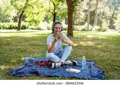 Young Woman Sitting In The Park Surfing The Song Play List In Her Phone And Listening The Music On The Wireless Headphones