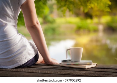 Young woman sitting and put down her hand beside coffee cup and notebooks on rustic wood bench with rural lake view in morning time on weekend