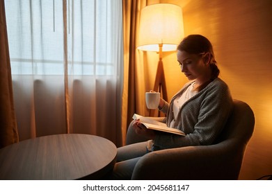 A Young Woman Is Sitting In A Room With A Cup Of Coffee. She Is Reading A Book. Home Interior, Light From A Floor Lamp. Rest, Relaxation, Home Comfort, Hotel. Quarantine.