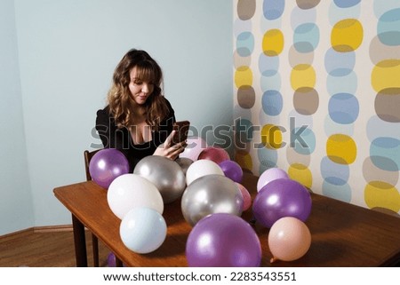 Young Woman Sitting at Table with Balloons and Looking at Phone, Against Colorful Background