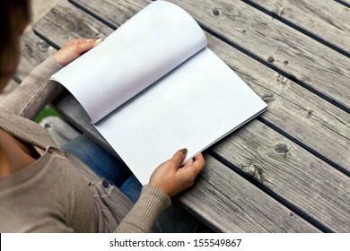 Young woman sitting at the table with a booklet with white pages. The white pages can be used for any logos, label signs or any graphic additions.