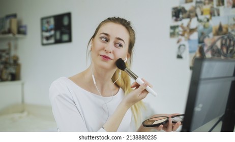 A young woman is sitting at a table in front of a mirror, listening to music with headphones and doing makeup