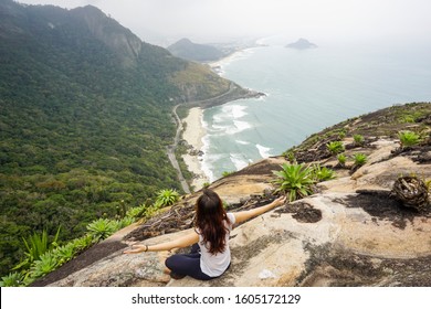Young woman sitting at the top of a hill with arms open looking at a wild beach in the middle of the forest, Rio de Janeiro, Brazil