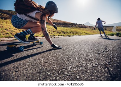 Young woman skating with her friend in background. Young people longboarding on rural road on summer day.