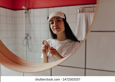 Young woman in sleep mask and white t-shirt looks in mirror and holds cream - Shutterstock ID 1472503337