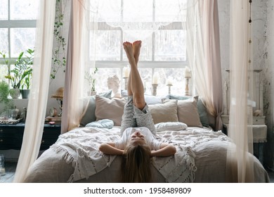Young woman in sleepwear raised legs up lying on comfortable bed in cosy bedroom with light bohemian interior. Careless female spending morning at home, enjoying lazy weekend