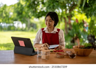 Young woman is slicing vegetables while following an online recipe on a digital tablet in a bright outdoor kitchen - Powered by Shutterstock