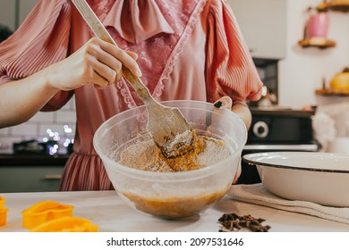 Young woman in smart dress kneads dough in kitchen with wooden spatula for Christmas gingerbread on Christmas eve, on blurred background of kitchen decorated with colorful lights. Making holiday baked