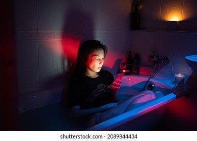Young woman with smartphone sitting in bathtub full of water in dark bathroom with neon light and burning candles, suffering from phone addiction disorder or nomophobia, addicted to social media